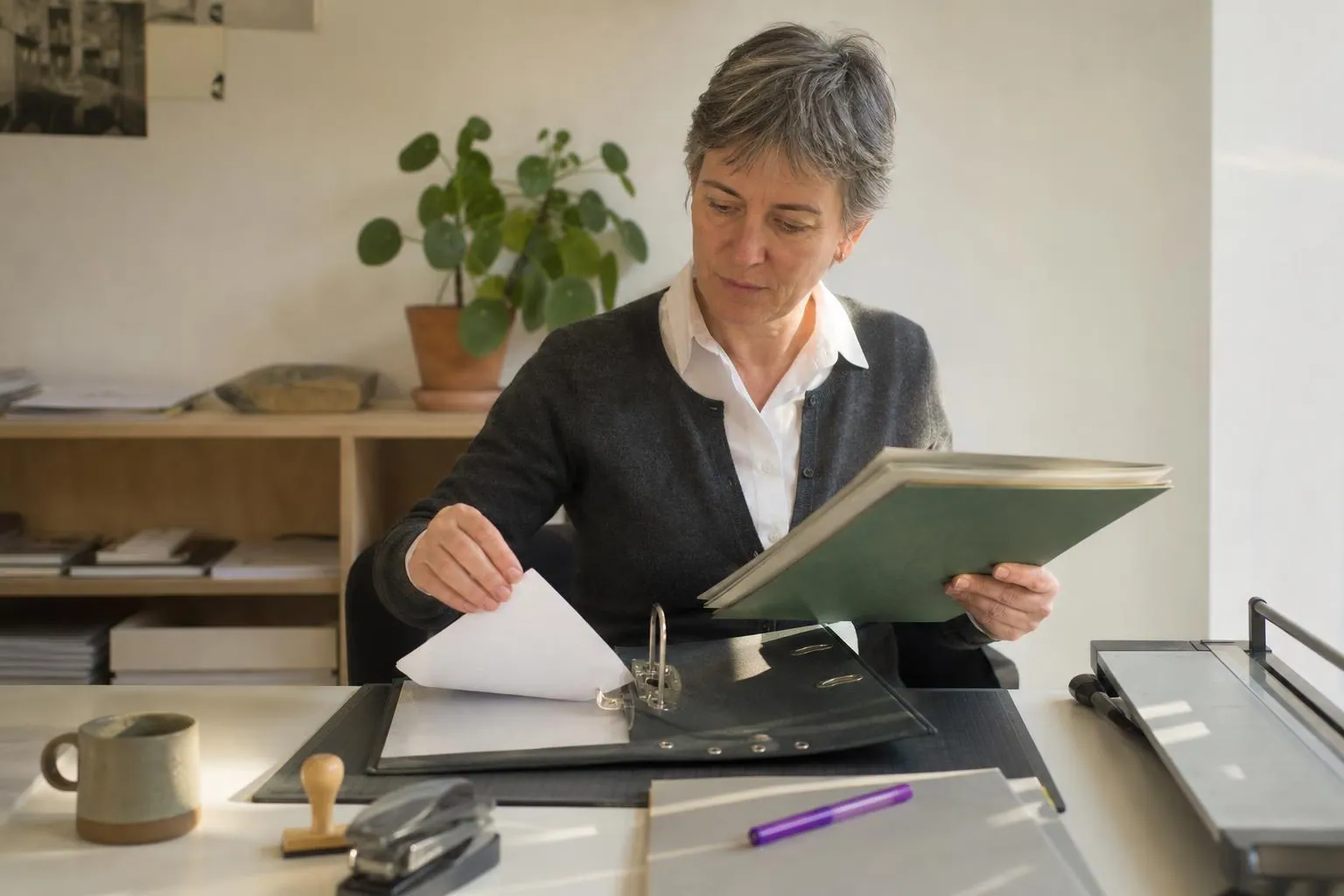 Une maître d’œuvre classe des attestations administratives dans un classeur à levier posé sur un bureau d’agence, en fin d’après midi.